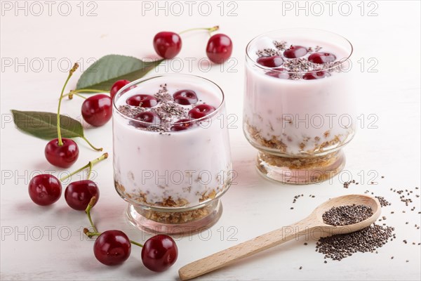 Yoghurt with cherries, chia seeds and granola in glass with wooden spoon on white wooden background. side view, close up