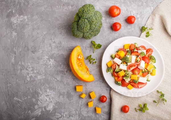 Vegetarian salad with broccoli, tomatoes, feta cheese, and pumpkin on white ceramic plate on a gray concrete background and linen textile, top view, copy space, flat lay