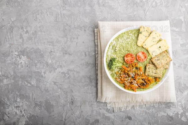 Green broccoli cream soup with crackers and cheese in white bowl on a gray concrete background and linen napkin. top view, flat lay, copy space