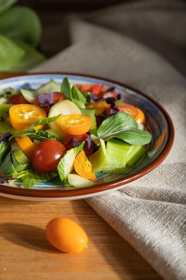 Vegetarian salad of pac choi cabbage, kiwi, tomatoes, kumquat, microgreen sprouts on a wooden background and linen textile. Hard light, contrast. Side view, close up, selective focus