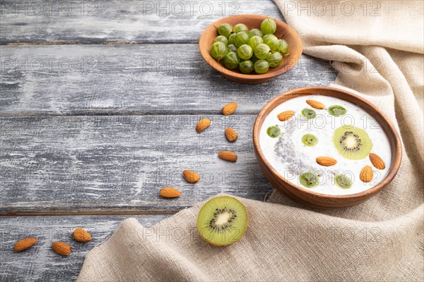 Yogurt with kiwi, gooseberry, chia and almonds in wooden bowl on gray wooden background and linen textile. Side view, copy space