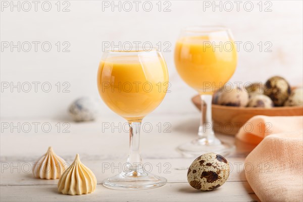 Sweet egg liqueur in glass with quail eggs and meringues on white wooden background. Side view, close up, high key