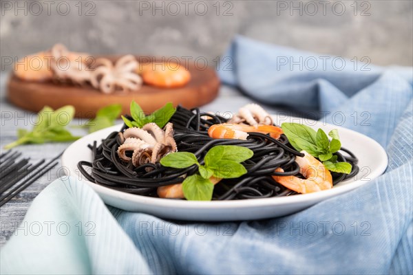 Black cuttlefish ink pasta with shrimps or prawns and small octopuses on gray wooden background and blue textile. Side view, close up, selective focus