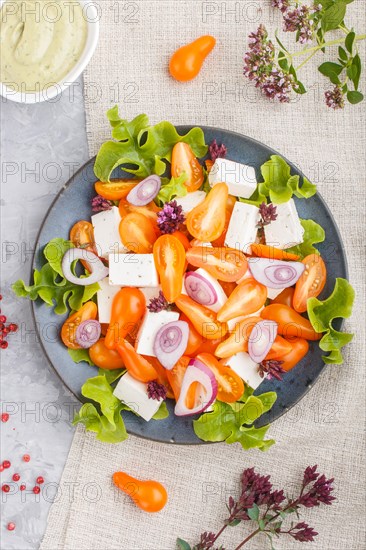 Vegetarian salad with fresh grape tomatoes, feta cheese, lettuce and onion on blue ceramic plate on gray concrete background and linen textile. top view, flat lay, close up