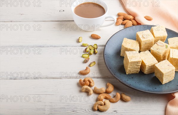 Traditional indian candy soan papdi in a blue ceramic plate with almond, pistache, cashew and a cup of coffee on a white wooden background with orange textile. side view, copy space
