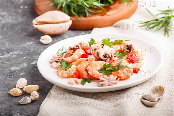 Boiled shrimps or prawns and small octopuses with herbs on white ceramic plate on a black concrete background and linen textile. side view, close up, selective focus