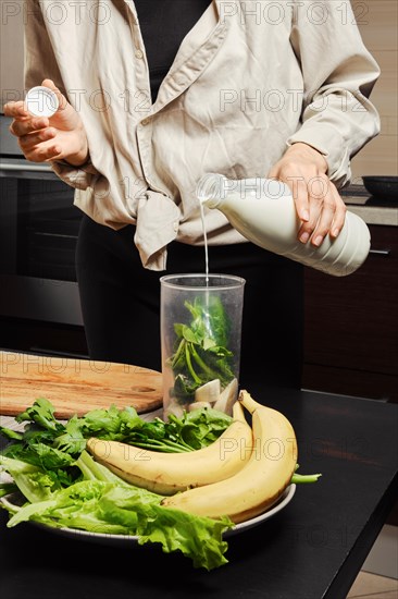Unrecognizable woman pouring milk into blender with banana and spinach