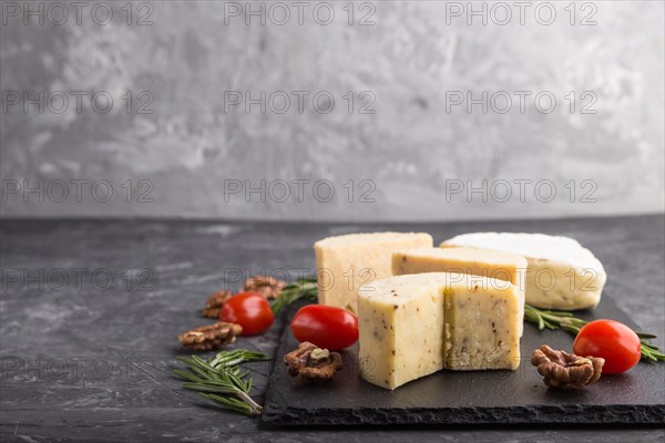 Cheddar cheese and various types of cheese with rosemary and tomatoes on black slate board on a black concrete background. Side view, copy space, selective focus
