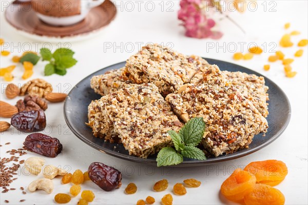 Homemade granola from oat flakes, dates, dried apricots, raisins, nuts in blue ceramic plate with a cup of coffee on a white wooden background. Side view, close up, selective focus
