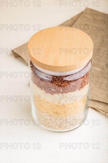Glass jar with different kinds of rice poured in layers on white background. side view, close up. Healthy food concept