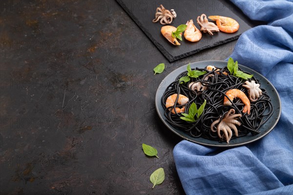 Black cuttlefish ink pasta with shrimps or prawns and small octopuses on black concrete background and blue textile. Side view, copy space
