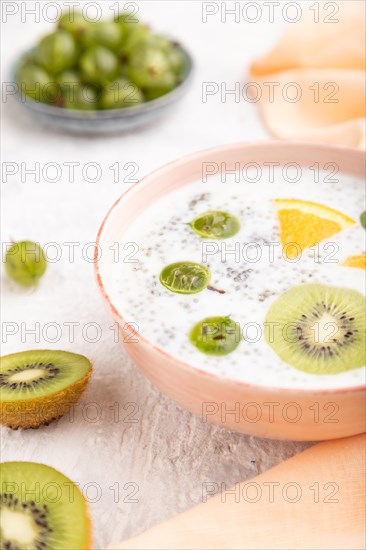 Yogurt with kiwi, gooseberry, chia in ceramic bowl on gray concrete background and orange linen textile. Side view, close up, selective focus