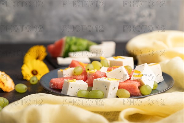 Vegetarian salad with watermelon, feta cheese, and grapes on blue ceramic plate on black concrete background and yellow linen textile. Side view, close up, selective focus