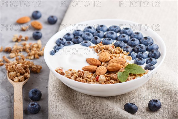 Yoghurt with blueberry, granola and almond in white plate with wooden spoon on gray concrete background and linen textile. side view, close up, selective focus