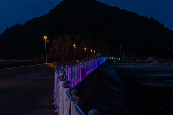 Night view of white metal fence on concrete pier lit with blue, purple and green LED lights in South Korea
