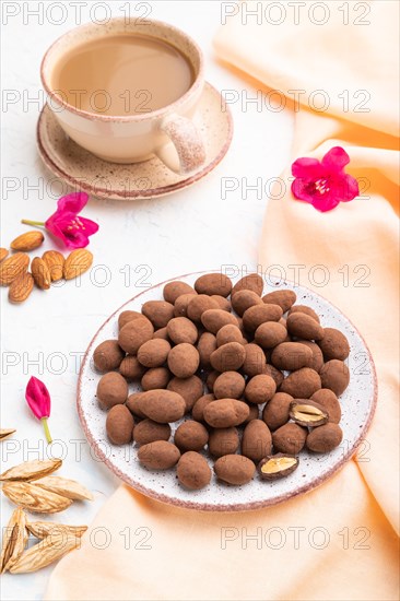 Almond in chocolate dragees on ceramic plate and a cup of coffee on white concrete background and orange linen textile. Side view, close up