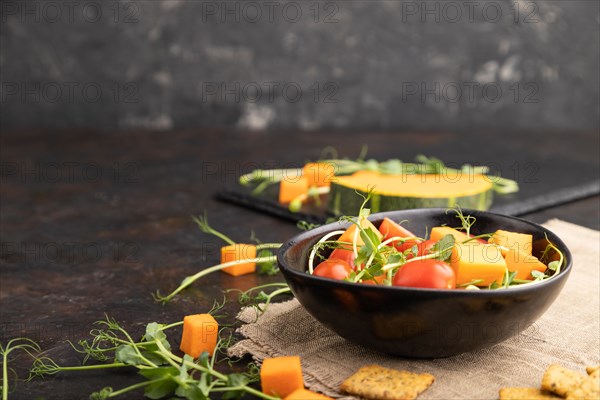 Vegetarian vegetable salad of tomatoes, pumpkin, microgreen pea sprouts on black concrete background and linen textile. Side view, copy space