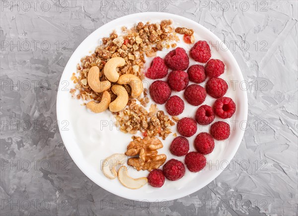 Yoghurt with raspberry, granola, cashew and walnut in white plate on gray concrete background and linen textile. top view, flat lay, close up