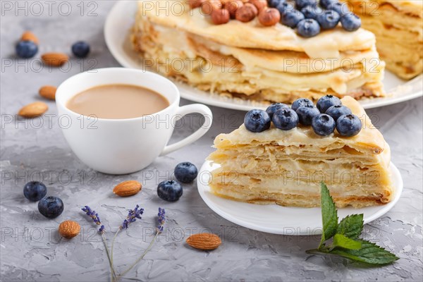 Homemade layered Napoleon cake with milk cream. Decorated with blueberry, almonds, walnuts, hazelnuts, mint on a gray concrete background and cup of coffee. side view, close up, selective focus