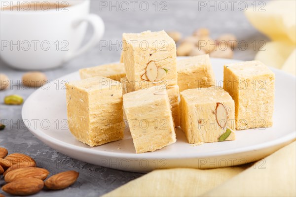 Traditional indian candy soan papdi in white plate with almond, pistache and a cup of coffee on a gray concrete background with yellow textile. side view, close up, selective focus