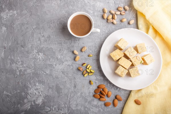 Traditional indian candy soan papdi in white plate with almond, pistache and a cup of coffee on a gray concrete background with yellow textile. top view, flat lay, copy space