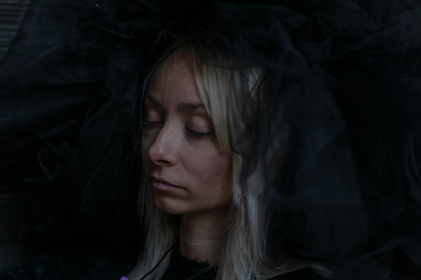 A young caucasian blonde woman's face is partially obscured by a dark veil, her eyes closed in a serene expression