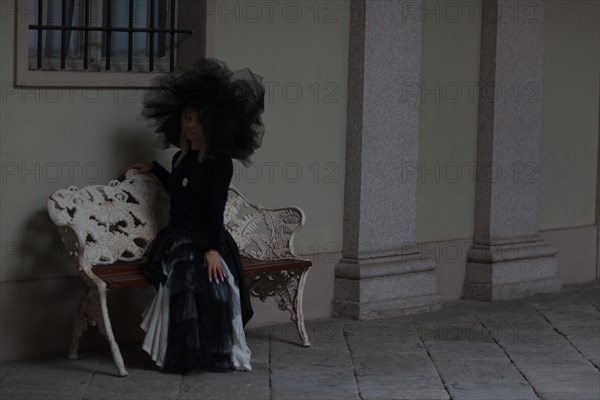 Thoughtful woman in black gothic attire sitting on a bench by a historical building, wear lace veil and hat, black and white dress