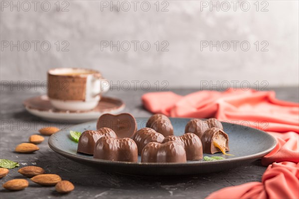 Chocolate caramel candies with almonds and a cup of coffee on a black concrete background and red textile. Side view, copy space