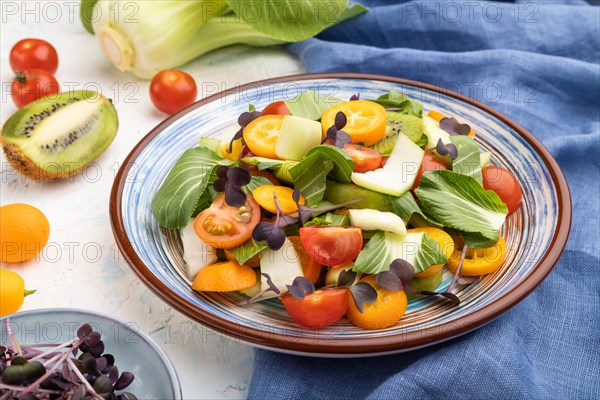 Vegetarian salad of pac choi cabbage, kiwi, tomatoes, kumquat, microgreen sprouts on a white concrete background and blue linen textile. Side view, close up