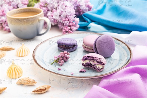 Purple macarons or macaroons cakes with cup of coffee on a white concrete background and magenta-blue textile. Side view, close up