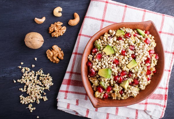 Salad of germinated buckwheat, avocado, walnut and pomegranate seeds in clay plate on black wooden background. Top view, flat lay