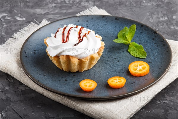 Cake with whipped egg cream on a blue ceramic plate with kumquat slices and mint leaves on a black concrete background with linen napkin. side view