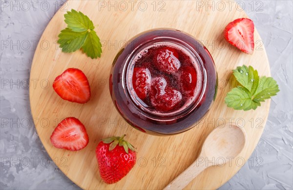 Strawberry jam in a glass jar with berries and leaves on gray concrete background. Homemade, top view, flat lay