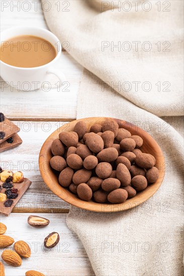 Almond in chocolate dragees in wooden plate and a cup of coffee on white wooden background and linen textile. Side view, close up