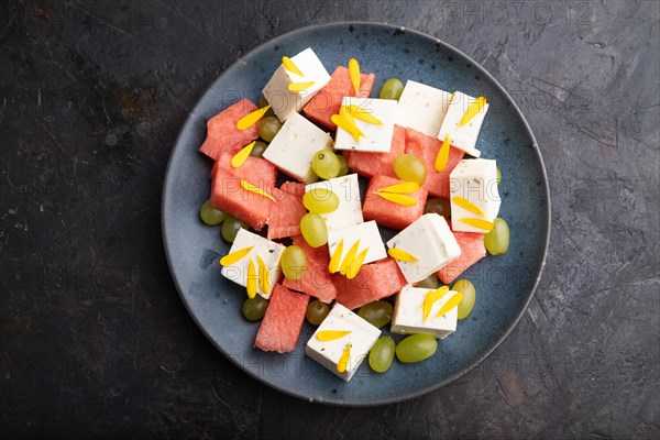 Vegetarian salad with watermelon, feta cheese, and grapes on blue ceramic plate on black concrete background. top view, close up, flat lay