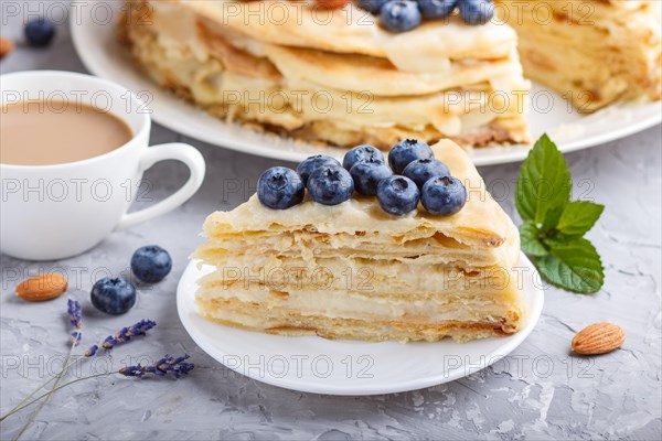 Homemade layered Napoleon cake with milk cream. Decorated with blueberry, almonds, walnuts, hazelnuts, mint on a gray concrete background and cup of coffee. side view, close up, selective focus