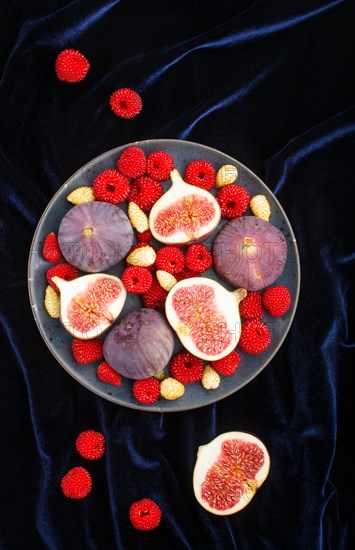 Fresh figs, strawberries and raspberries on blue ceramic plate on black concrete background and blue velvet textile. top view, flat lay, close up