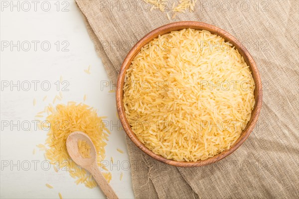 Wooden bowl with raw golden rice and wooden spoon on a white wooden background and linen textile. Top view, flat lay, close up