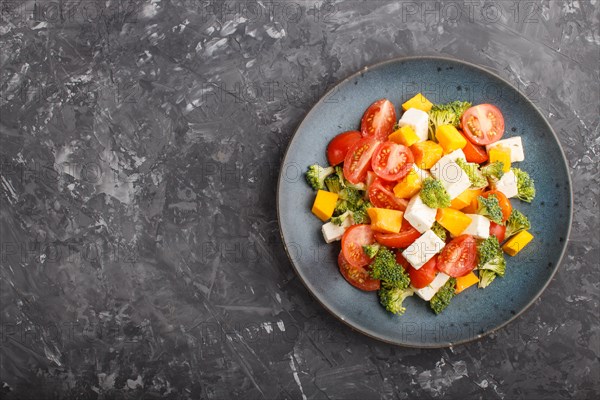 Vegetarian salad with broccoli, tomatoes, feta cheese, and pumpkin on a blue ceramic plate on a black concrete background, top view, copy space, flat lay