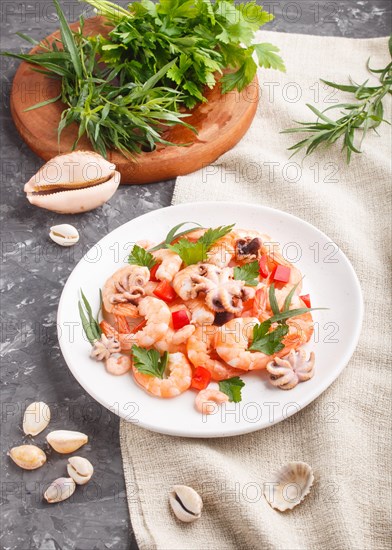 Boiled shrimps or prawns and small octopuses with herbs on white ceramic plate on a black concrete background and linen textile. side view, close up