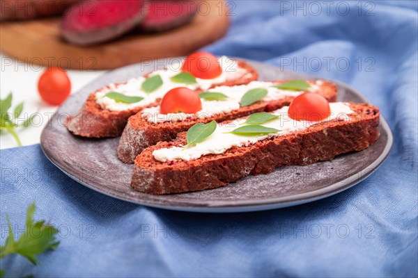 Red beet bread sandwiches with cream cheese and tomatoes on white concrete background and blue linen textile. Side view, selective focus, close up