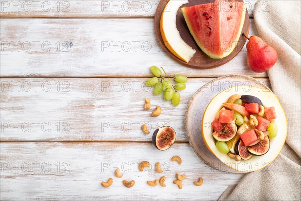 Vegetarian fruit salad of watermelon, grapes, figs, pear, orange, cashew on white wooden background and linen textile. Top view, flat lay, copy space