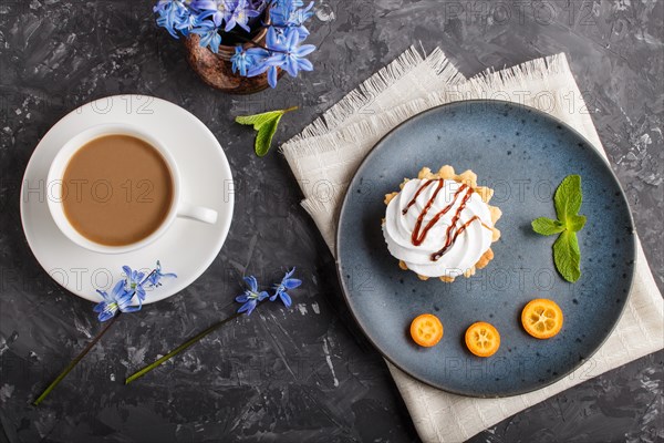 Cake with whipped egg cream on a blue ceramic plate with kumquat slices and mint leaves on a black concrete background with linen napkin and cup of coffee. decorated with blue flowers. flat lay, top view