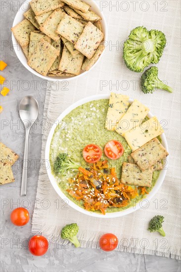 Green broccoli cream soup with crackers and cheese in white bowl on a gray concrete background and linen napkin. top view, flat lay, close up