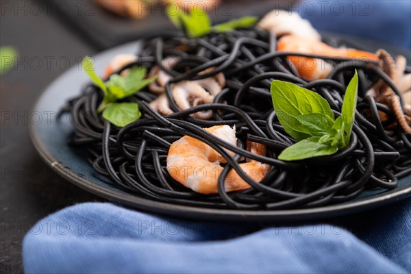 Black cuttlefish ink pasta with shrimps or prawns and small octopuses on black concrete background and blue textile. Side view, close up, selective focus