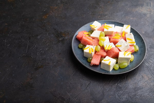 Vegetarian salad with watermelon, feta cheese, and grapes on blue ceramic plate on black concrete background. Side view, copy space