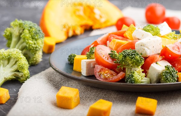 Vegetarian salad with broccoli, tomatoes, feta cheese, and pumpkin on a blue ceramic plate on a black concrete background and linen textile, side view, close up, selective focus