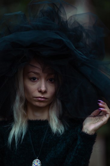 Mysterious white young blonde woman in a dark setting wear a large black tulle hat and veil and a black furry dress