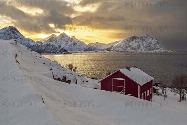 Ein rotes Haus vor einem winterlichen Sonnenuntergang mit verschneiten Bergen auf den Vesteralen