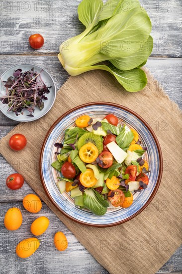 Vegetarian salad of pac choi cabbage, kiwi, tomatoes, kumquat, microgreen sprouts on gray wooden background and linen textile. Top view, flat lay, close up